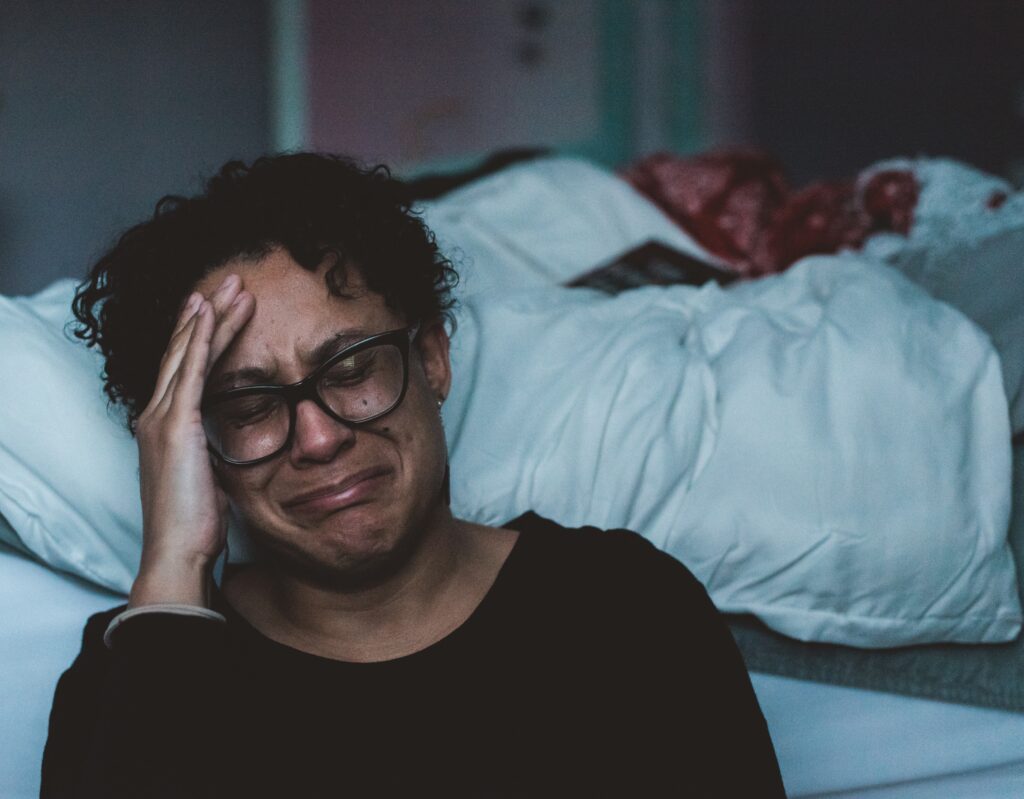 A woman of colour with short curly hair, wearing a pair of thick black frame glasses, is crying beside her undone bed.
