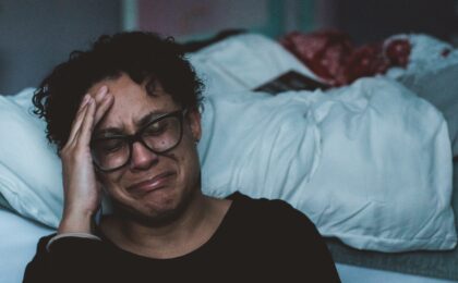 A woman of colour with short curly hair, wearing a pair of thick black frame glasses, is crying beside her undone bed.